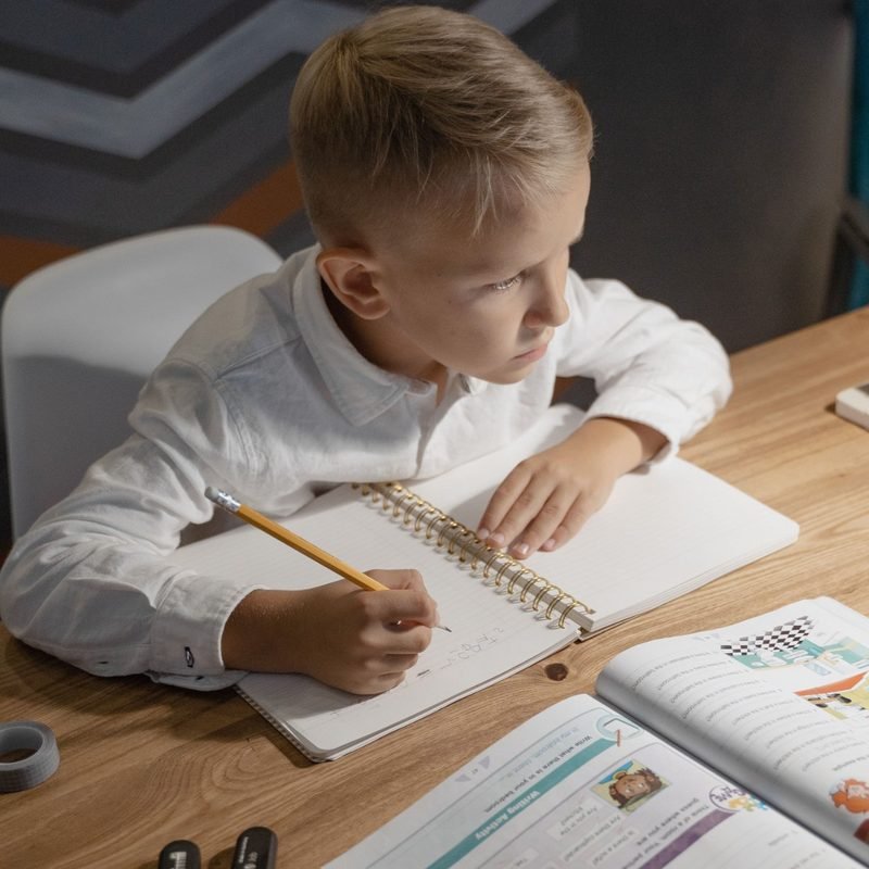 Primary school boy writing in a notebook at a desk with a tablet