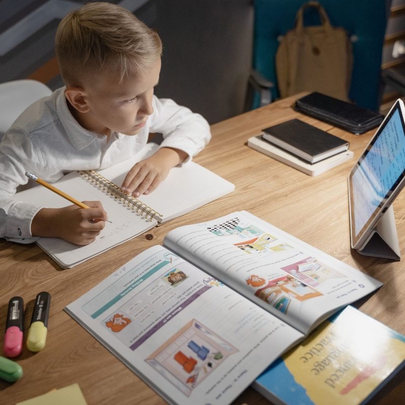 Primary school boy writing in a notebook at a desk next to a tablet